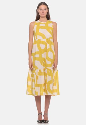 Donna in piedi, dritta, che indossa un vestito midi bianco senza maniche con grandi motivi astratti gialli e tacchi neri con punta affilata.