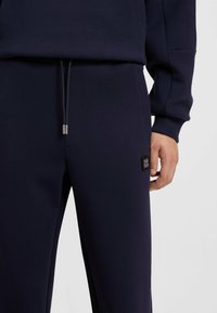 Los pantalones de chándal azul marino presentan una cintura con cordón, bolsillos laterales y una tela texturizada. Un parche de la marca está situado en la cadera izquierda.
