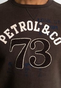 Tmavě hnědá mikina s bílým vyšívaným nápisem "PETROL & CO" a texturovaným číslem "73". Měkká tkanina s kulatým výstřihem a jemnými tiskovými detaily.