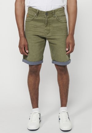 Shorts in denim verde oliva con risvolti, tasche frontali, cuciture tono su tono e una patch con logo in vita. Abbinati a sneaker bianche.