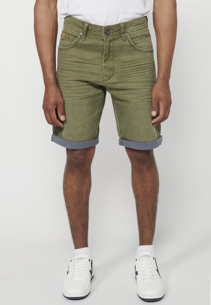 Shorts de denim verde oliva con dobladillos enrollados, bolsillos frontales, costuras tonales y una etiqueta de marca en la cintura. Combinados con zapatillas blancas.