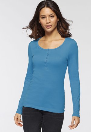 Langarmshirt mit Rundhalsausschnitt in einfarbig Blau, mit Knopfleiste und glatter Textur. Kombiniert mit dunklen Jeans.