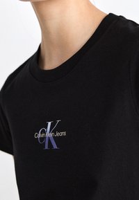 Person, der ein schwarzes T-Shirt mit Rundhalsausschnitt trägt, auf dessen Brust sich ein kleines "Calvin Klein Jeans"-Logo in Blau und Weiß befindet.