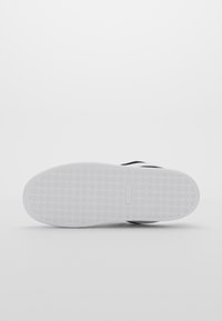Semelle d'une basket avec une semelle extérieure en caoutchouc blanc présentant un petit motif en grille et une empreinte rectangulaire près du centre.