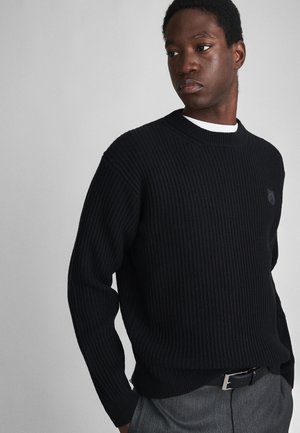 Black ribbed sweater with a crew neckline, textured fabric, and subtle shoulder seams, paired with gray trousers and a black belt.