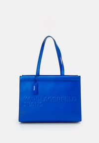 Sininen kantokassi, joka on valmistettu sileästä materiaalista. Sen etupuolella on kohokuvioitu "KARL LAGERFELD JEANS" -teksti ja pieni merkki. Kaksinkertaiset tukevat kahvat helpottavat kantamista.