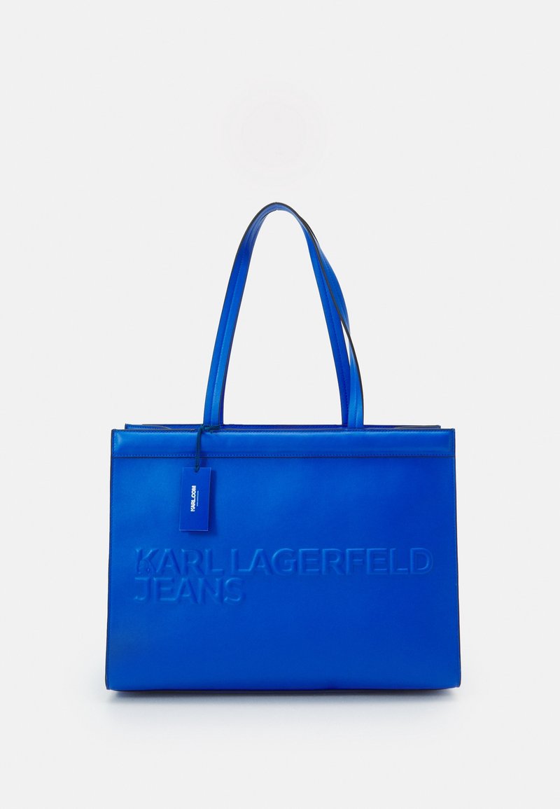 Sininen kantokassi, joka on valmistettu sileästä materiaalista. Sen etupuolella on kohokuvioitu "KARL LAGERFELD JEANS" -teksti ja pieni merkki. Kaksinkertaiset tukevat kahvat helpottavat kantamista.