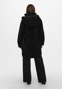 Abrigo de forro polar negro con capucha, diseño oversize, tejido texturizado y mangas largas. Lleva puesto sobre pantalones negros de corte ancho.