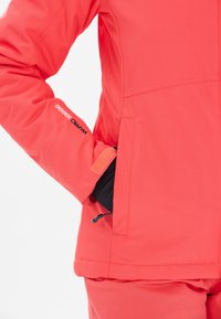 Helle Koralljacke mit weicher Textur, ausgestattet mit einer Reißverschlusstasche, einem Klettverschluss zur Anpassung der Ärmelbündchen und einem Logo auf dem Ärmel.