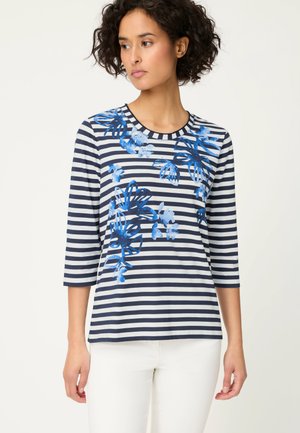 Kobieta ubrana w granatowo-białą koszulę w paski z niebieskimi kwiatowymi nadrukami, o długości rękawów 3/4, w połączeniu z białymi spodniami, stojąca na jednolitym tle.