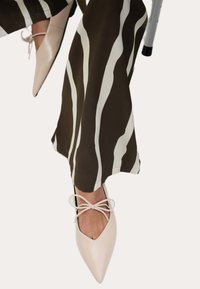 Personne portant un pantalon rayé marron et blanc et des ballerines beiges pointues attachées avec des nœuds, assise sur une chaise.