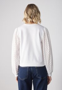 Pullover bianco con maniche in pizzo ad occhio di pernice testurizzato e orlo aderente. Presenta un collo rotondo e polsini elasticizzati. Jeans di denim visibili sotto.
