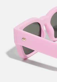 Occhiali da sole rosa con una montatura in plastica spessa, lenti scure e un piccolo accento metallico dorato sul lato. Texture liscia e forma audace.