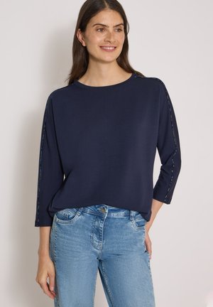 Femme aux longs cheveux bruns portant un haut bleu marine à manches longues et un jean bleu clair, souriante et debout devant un fond uni.