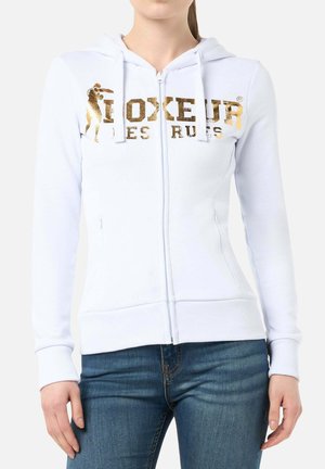 White zip-up hoodie with gold boxing-themed logo across the chest, worn by a person in blue jeans, shown from neck to hips.
