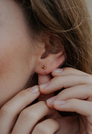 Zlatý náušnicový kôstok s trblietavým zoskupením troch guľovitých prvkov, zobrazený na uchu modelky s rukami jemne dotýkajúcimi sa náušnice.