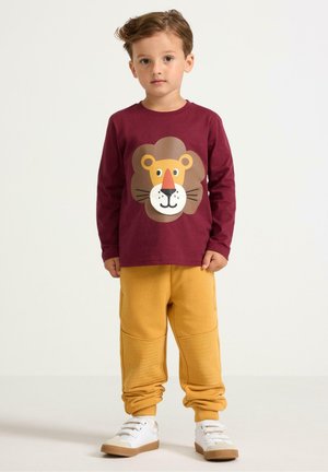 Camicia a maniche lunghe bordeaux con una grafica di leone, abbinata a pantaloni felpati gialli con dettagli testurizzati e sneakers bianche con suola marrone.
