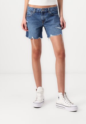 Person iført blå denim cut-off shorts med mellemhøjt talje og hvide high-top sneakers, stående mod en ensfarvet hvid baggrund.