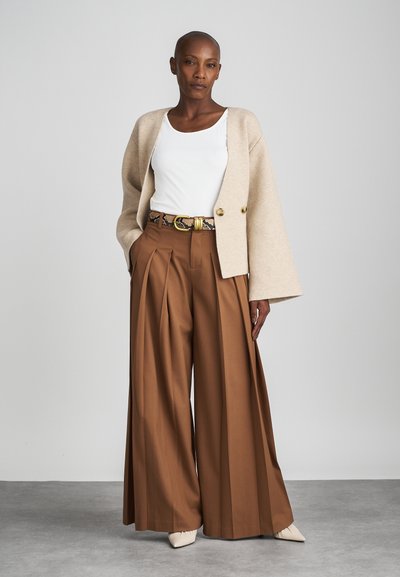 Woman wearing beige cropped cardigan over white top, high-waisted wide brown trousers with snakeskin belt, and pointed cream shoes, standing.