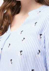 Blouse rayée bleu et blanc avec un décolleté en V, ornée de petits motifs de pingouins en noir et blanc, et d'une texture de tissu douce et légère.