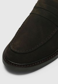 Mocassin en daim marron foncé avec des détails de couture et des accents élastiques. Bout arrondi et semelle en caoutchouc profilée basse pour le confort.