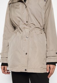 Béžová, lehká bunda s staženým pasem, patentkami a bočními kapsami. Má hladkou texturu a nastavitelné manžety.