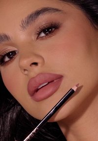 Ołówek do ust z ostrym końcem, trzymany blisko pełnych ust, w kolorze mauve, o gładkiej teksturze, czarne opakowanie z metalicznymi akcentami, w naturalnym makijażu.