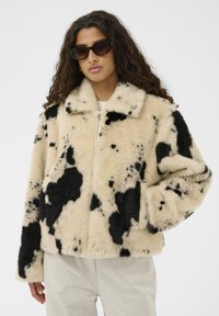 Jakna iz lažnega krzna z bež in črnim vzorcem krave, z zadrgo, ovratnikom, širokim krojem in mehko teksturo. Model nosi sončna očala in svetle hlače.