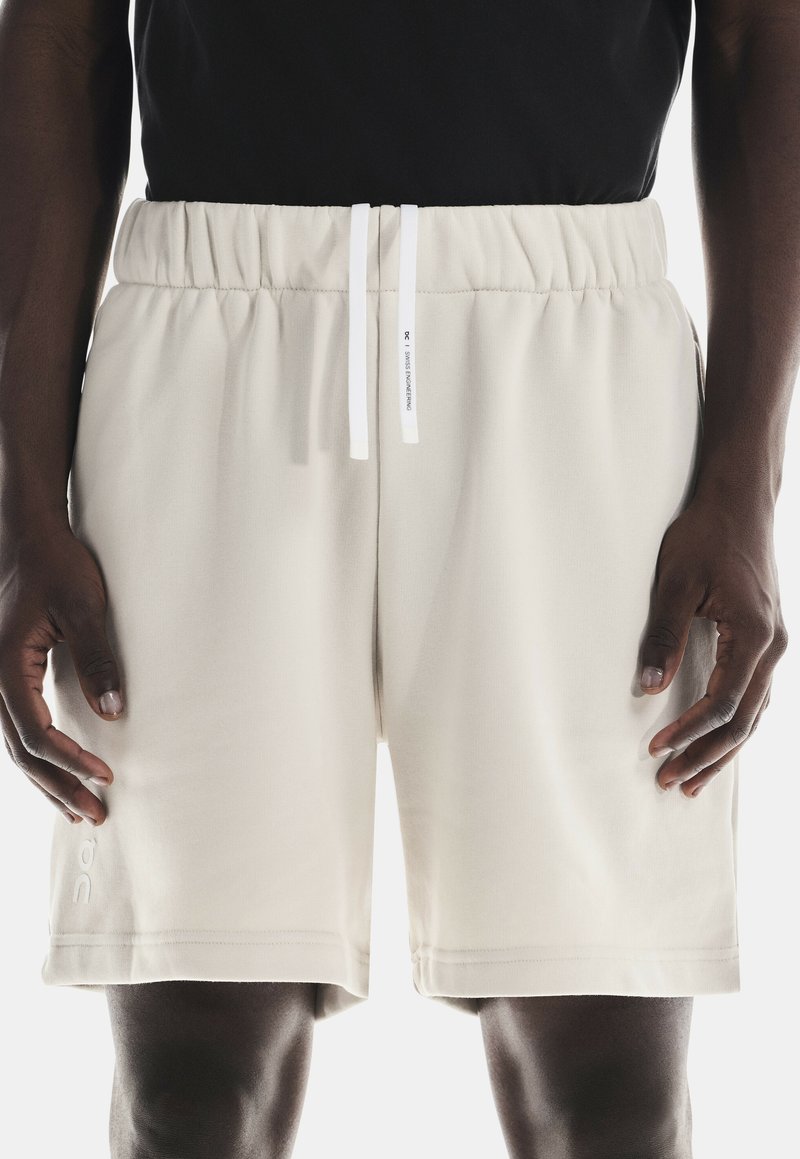 Beige Sportshorts mit einem elastischen Bund, weißen Zugbändern und einem Logo am linken Bein. Weicher, glatter Stoff mit lockerer Passform.