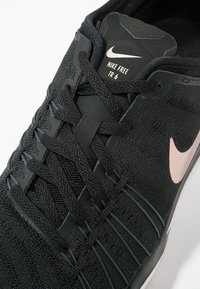 Musta Nike Free TR 6 -kengät, joissa on tekstuuripintainen päällinen, litteät nauhat ja vaaleanpunaiset Swoosh-yksityiskohdat. Suunniteltu joustavalla pohjalla.
