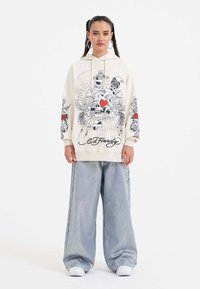 Mikina s kapucí v krémové barvě s grafickým potiskem, na kterém je lebka a růže; v kombinaci s lesklými světle modrými širokými kalhotami a bílými teniskami.