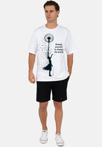 Witte katoenen T-shirt met een zwarte paardenbloem en silhouet graphic met de tekst "Verander jezelf om de wereld te veranderen." Gedragen met zwarte korte broek.