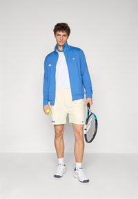 Jachetă sportivă albastră cu guler înalt, tricou alb, şorturi crem deschis, ținând o minge de tenis și o rachetă, purtând adidași albi.