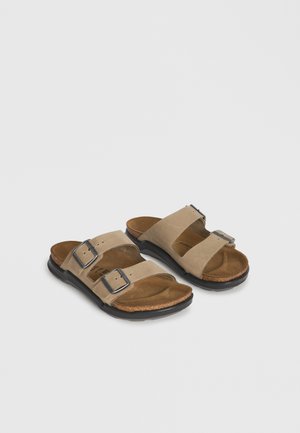 Beige sandaler med to stropper med justerbare metallspenner, formede korkfotsenger og svarte gummisåler på ensfarget bakgrunn.
