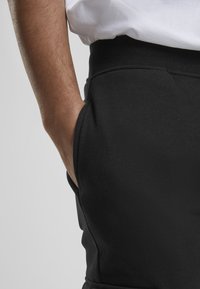 Black sweatpants with a smooth texture, featuring side pockets and a fitted waistband, shown with a hand placed in the pocket.