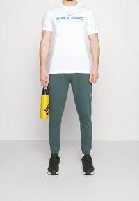 Vit Nike Pro T-shirt med blå logotyp, gröna joggers, svarta sneakers och en gul vattenflaska med svart Nike-logotyp.