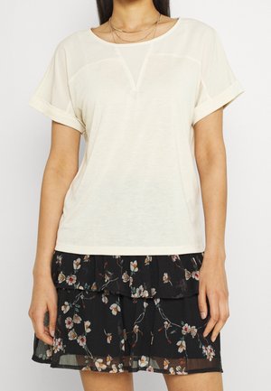 T-shirt crème clair à manches courtes roulées, arborant une texture lisse et un élément de design en V au niveau de l'encolure, accompagné d'une jupe noire à motifs floraux.