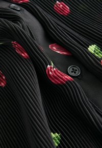 Tissu noir plissé avec des motifs floraux rouges, comportant un détail de bouton noir. La texture met en valeur des rainures verticales qui rehaussent le design.