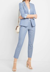 Světle modrý upravený oblek s úzkým střihem, se vzorovanými manžetami a v kombinaci s béžovými podpatky. Doplněno malou kabelkou přes rameno.