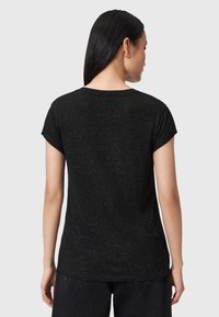 Mujer con cabello largo y negro que lleva una camiseta de manga corta negra con manchas, de espaldas, de pie contra un fondo claro y liso.