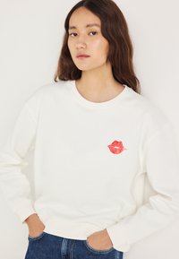 Felpa bianca con scollo rotondo, caratterizzata da una grafica di labbra rosse sul petto. Tessuto morbido e vestibilità rilassata. Abbinata a jeans blu.