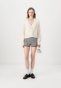 Creme cardigan med v-udskæring og knaplukning, parret med sort-hvide stribede shorts. Modellen holder en lille, lyserød håndtaske og bærer sorte sko med hvide sokker.