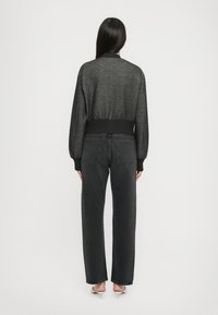 Blouson bomber noir en tricot avec ourlet et poignets côtelés, porté sur un jean noir droit. Texture lisse et légère variation de couleur visible.