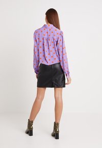 Blusa lunga viola a maniche lunghe con pois arancioni, dettaglio di piega sul retro e gonna in pelle nera. La modella indossa stivaletti con tacco verde oliva.