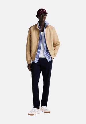Bomber jacket beige chiaro, camicia a righe blu e bianche, canotta bianca, pantaloni blu scuro e sneakers bianche con suole in gomma. Dettagli visibili del cappellino bordeaux.