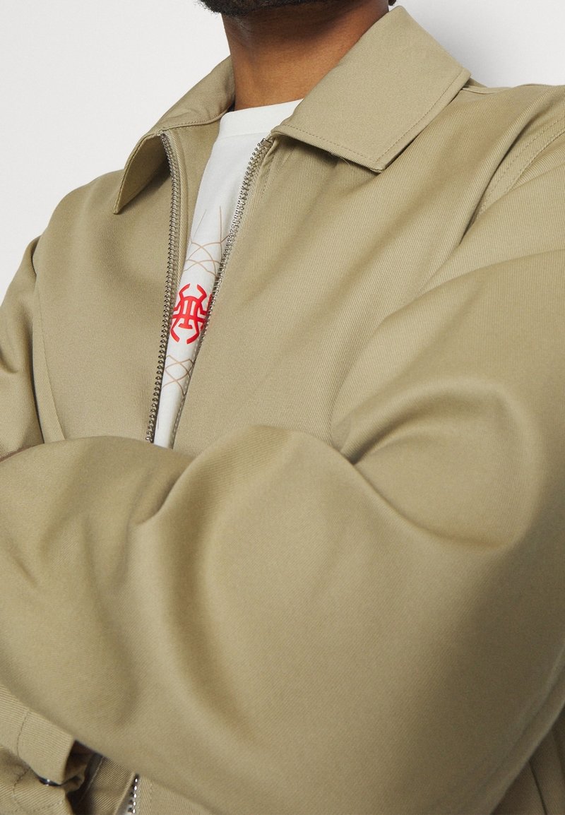 Vaaleanruskea vetoketjullinen takki, jossa on klassinen kaulus ja sileä, teksturoitu pinta. Sen päällä on graafinen valkoinen t-paita, jossa on punaisia kuvioita.