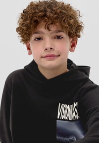 Czarna bluza z kapturem z napisem "VISIONMASK" w wyraźnych białych literach, krótki krój oraz graficzny nadruk po lewej stronie.