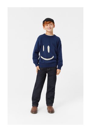 Maglione in maglia blu navy con design di faccina sorridente bianca, abbinato a jeans scuri e stivali marroni. La vestibilità è rilassata con scollo rotondo e orlo a coste.