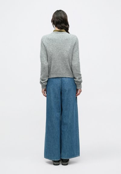 Άτομο που στέκεται με την πλάτη στην κάμερα, φοράει γκρι πουλόβερ, μπλε τζιν παντελόνι με φαρδιά μπατζάκια και μαύρα παπούτσια, σε απλό λευκό φόντο.