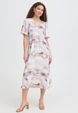 Femme aux cheveux bouclés portant une robe midi à manches courtes, col en V, motif marbré et des sandales beiges à talons blocs, debout sur fond blanc.